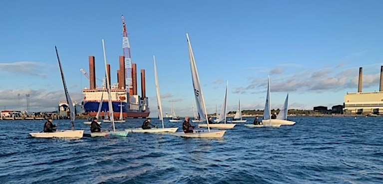 The East Antrim Boat Club dinghy fleet prepare for a start at Larne Harbour and in the background (left) is MPI Resolution, the world's first purpose-built vessel for installing offshore wind turbines currently based in Larne