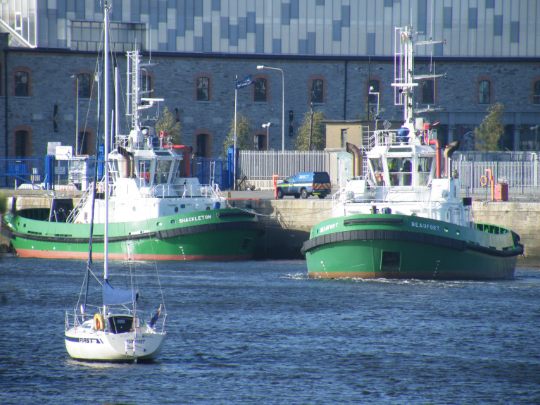 Towage Twins Relocate: Dublin Port owned tugs, Shackleton berthed on left while Beaufort sets off on towage duties from their previous station berth located on the North Quay Wall Extension next to the Tom Clarke Bridge (East-Link) not in shot but the Three Arena is seen above. Now the tugs have a more centrally located station berth downriver to faciliate easier operations and for the safety of crew using a custom built pontoon. Note, the tugs don't feature funnel's, instead use exhaust (pipes) uptakes positioned on both sides of the tug master's wheelhouse. 